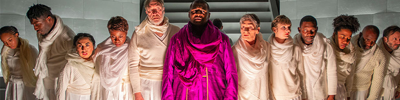 A group of people stand onstage in white robes. In the center, there is a man wearing a bright violet robe. 