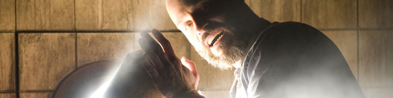 A man kneels in front of a wall and holds his hand over a portal with light streaming out. 