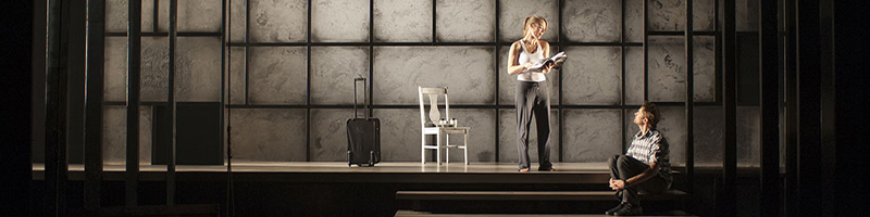 A woman stands in front of a chair, holding a book in her hand. A man is seated on stairs in front of her. 