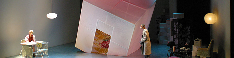 A large white cube on an angle sits onstage. There is someone at a table in the corner and someone standing across from them trying to get their attention.