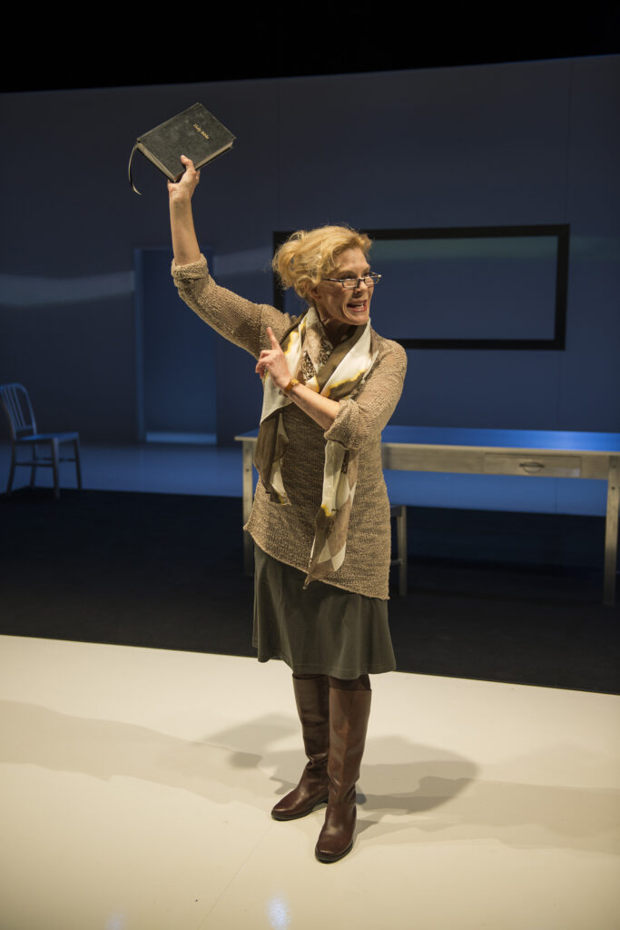 A woman stands onstage holding a book overhead and pointing to it.