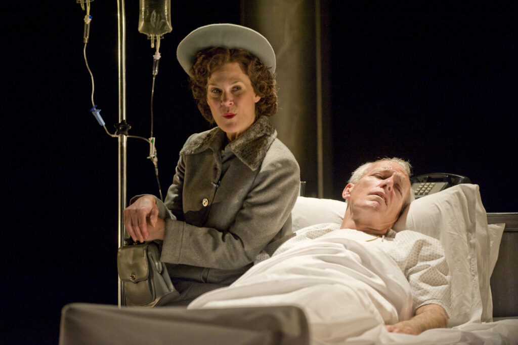 A woman wears a fancy coat and sits next to someone on a hospital bed.