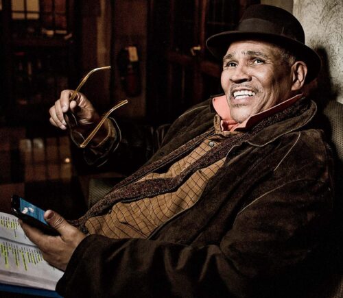 Ron OJ Parson holding his glasses, a script, and a phone; he is smiling.