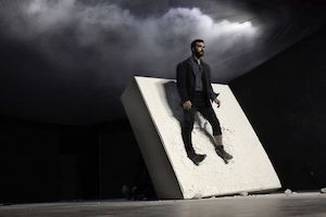 A black stage with a white light and smoke in the top right corner; downstage, a white slab tilts forward at a steep angle and a man leans against it.