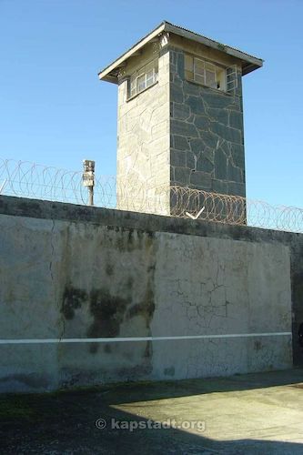 A blue sky with a gray wall in front, with barbed wire; a large tower rises up from the wall.