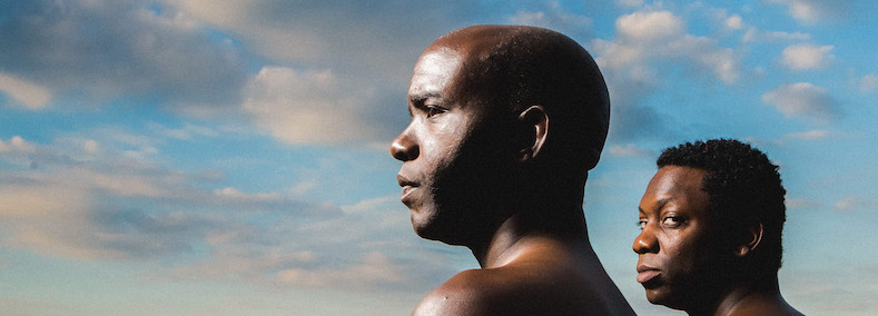 A blue sky with clouds in the background, as one man looks to his left, while another man looks directly at the camera.