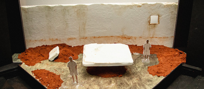 A model of a white wall; red sand against the wall and curving around front; a large, white slab, and two cut-out male figurines.