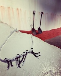 A necklace made of rusty iron nails on a white slab, with red sand, white walls, and two shovels in the background.