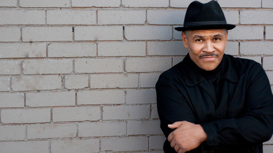 A man in a black turtleneck and black hat stands against a gray brick wall. 