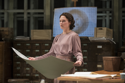 A woman in a pale pink dress holds a large sheet of paper with a projection behind her.