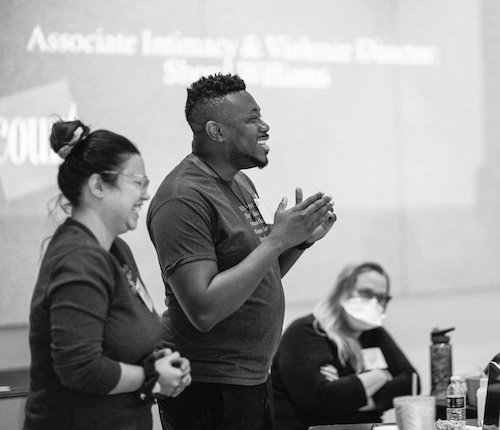 A black-and-white photo of a man and a woman standing side-by-side, giving a presentation. The photo is captured in profile.