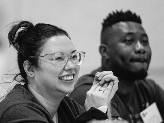 A black and white photo of a man and woman sitting next to one another listening intently; the woman is smiling.