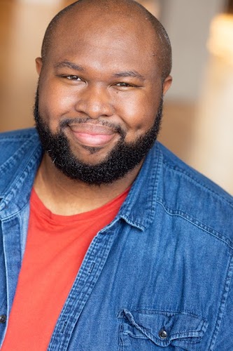 A bald man with a beard wears a red t-shirt beneath an open, denim button-up.