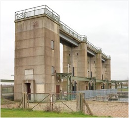 A tall, light brown structure with three columns and a bridge/walkway connecting each tower.