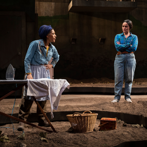 A woman irons in a field as a woman in working clothes stands behind her, with her arms crossed. The woman in work clothes looks inquisitive, the woman ironing looks irritated.