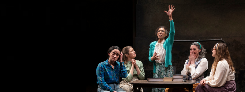 A group of women sit at a table with books. One woman stands in the center of the group with her hand raised to the ceiling; the other three women look at her with love, respect, and admiration. The woman seated at the far left of the group looks bored.