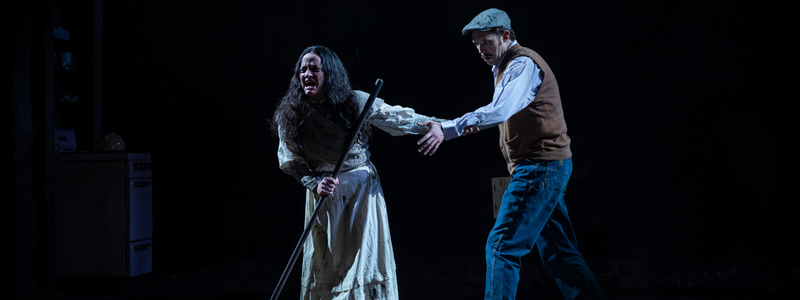 A woman in a tattered, white dress holds a gardening hoe and screams, while she reaches back and grasps the arm of a man who looks terrified.