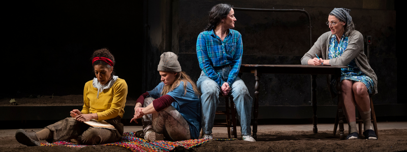 Two women dressed as children sit on the floor - one is coloring, one is pouting. A woman sits in a chair above them, and she is seated across from an older woman wearing a bandana. This woman and the older woman are in conversation.