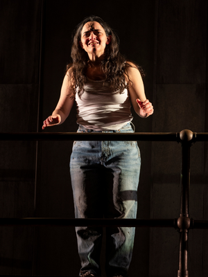 A woman in a white tank top and blue jeans stands on a platform with her hands raised slightly.