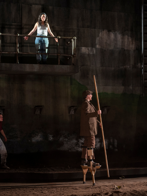 A woman in a brown suit walks on stilts; there is a woman on a lighted platform above her wearing a white tank top and jeans.