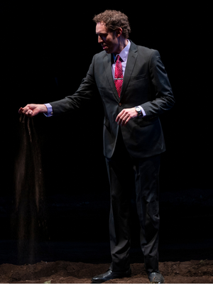 A man in a suit holds dirt in his right hand and slowly lets it trickle down to the ground.