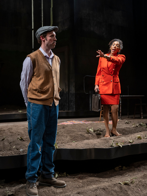 A man in a light colored button up and a beige sweater vest stands in a field, as a woman in an orange suit talks to him with her arm outstretched.