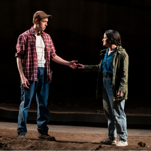 A man and a woman - both wearing work clothes and both looking pensive - reach out for each others' hands.