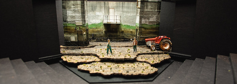 A black box with tiers to mimic stairs. In the center there is a small model of the set for Caryl Churchill's FEN. There are walls with ombre (brown to green) stains, tiered layers of dirt on the ground, a tractor, and two small figurines.