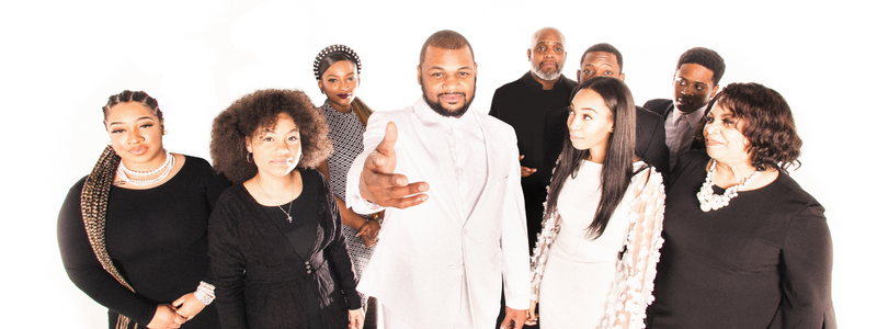 A man in a white suit holds out his hand to the camera; he is surrounded by people in black clothes.