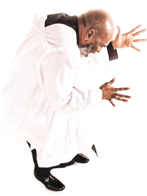 A man in white robes with black underclothes is bent over with his hands stretched out in front of him.