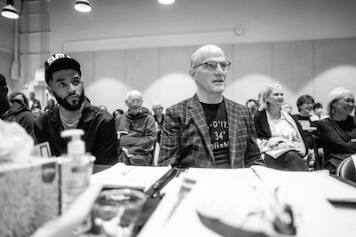 Two men sit at a table covered in scripts, pens, tissue boxes, and hand sanitizer. They are both looking to their left, focused.