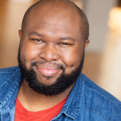 A bald man wearing a denim shirt smiles at the camera.