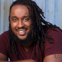 A man wearing a maroon shirt is leaning on his arms that are crossed in front of him. He is smiling at the camera.