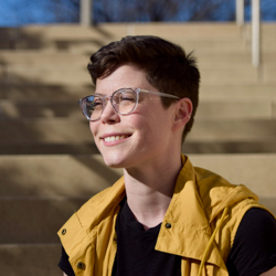 A woman wears a black t-shirt and a yellow vest. She has short hair and glasses, and is smiling as she looks in the distance.