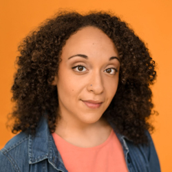 A woman is wearing a peach shirt with a denim shirt over that. She is smiling at the camera.