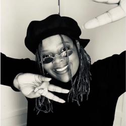 A woman wearing a black turtleneck and black beret smiles and holds up peace signs to the camera.