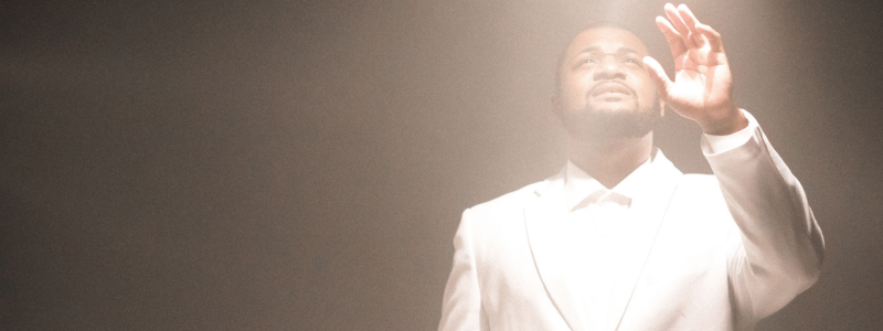 A man in a white suit stands against a gray background. He is bathed in warm light, and is raising his eyes and his left hand up to the ceiling. He looks inquisitive.