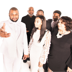 A man in a white suit stands with his arm outstretched to the viewer. There is a woman in a white dress to the right, and a group standing behind them dressed in black.