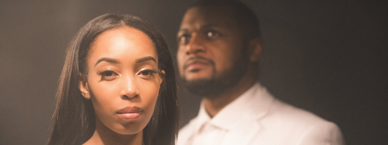 A woman is looking straight at the camera. Behind her, a man in a white suit looks off-camera. The background is blurry, and there is a light source above the two people.