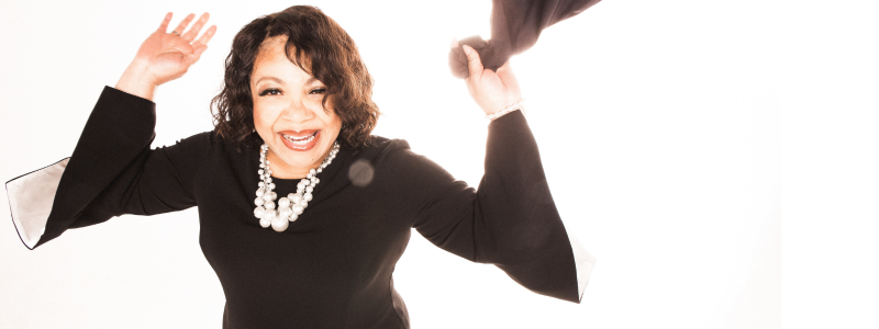 A woman in a black dress and a pearl necklace holds her hands in the air; she is smiling. In her left hand, she is holding a piece of black fabric that she's waving in the air.