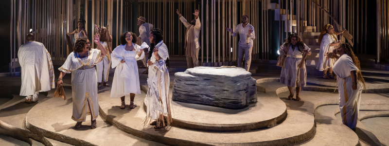 A group of people in white, beige, and gold clothes sing around a low, flat, gray rock.
