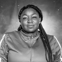 A woman wearing braids and a sports jacket looks at the camera and smiles.