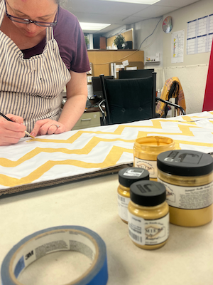 A woman paints gold zig-zags onto white fabric.