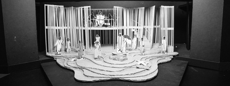 A set model that has a series of wooden slats that comprise undulating walls. There are shallow stone steps leading to a rock in the center of the stage, with paper figurines representing the cast scattered across the set.