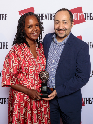 A woman in a red dress stands next to a man in a blue suit. They're smiling and holding a Tony Award.