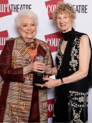 A woman in a red and orange patterned coat stands next to a woman in black formalwear. They're both smiling and holding a Tony Award.