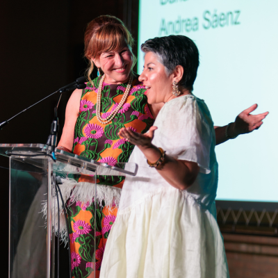 Two women deliver remarks at a podium. One is in a colorful patterned outfit - she is grinning and holding her arm out behind the other woman. This woman is wearing a white dress and speaking at a podium. Both look happy.