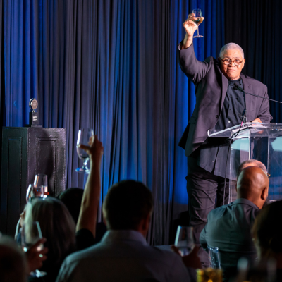 A man stands at a clear podium onstage and raises a glass in a toast.