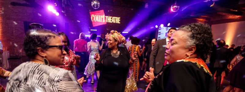 Guests dance on a dance floor; there are disco balls hanging from the ceiling above them and there's a projection that says "Court Theatre" on the far wall.