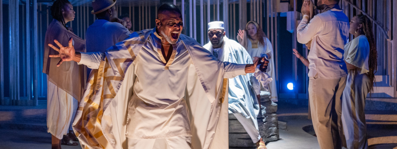 A man in a white outfit wears a white and gold cape. He is bending his knees, squinting his eyes, and spreading his arms - he looks as if he is shouting or singing passionately. There is a group of people singing behind him.